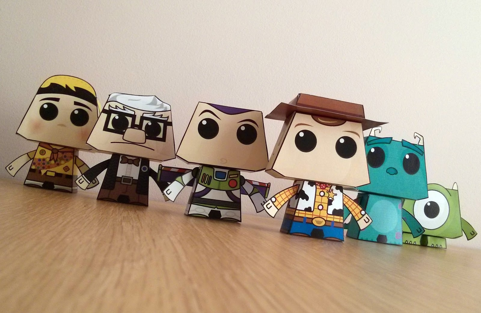 Seis brinquedos artesanais de papel montados em cubo, com personagens coloridos, apoiados em uma superfície de madeira